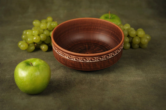 Handmade rustic clay bowl with decorative pattern, surrounded by green apples and grapes.