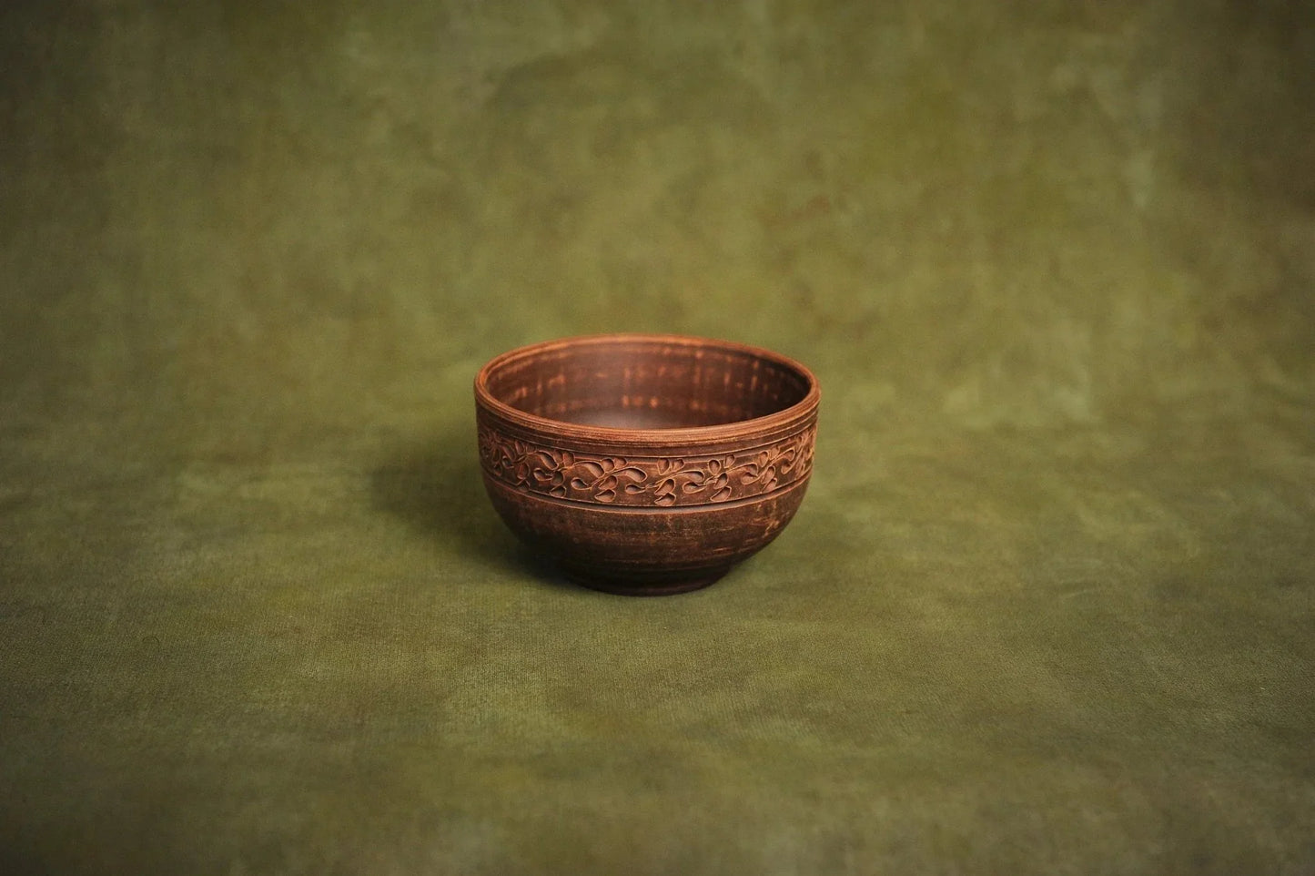 Handmade deep red clay ceramic bowl with ornate rim on a green background, Ukrainian pottery