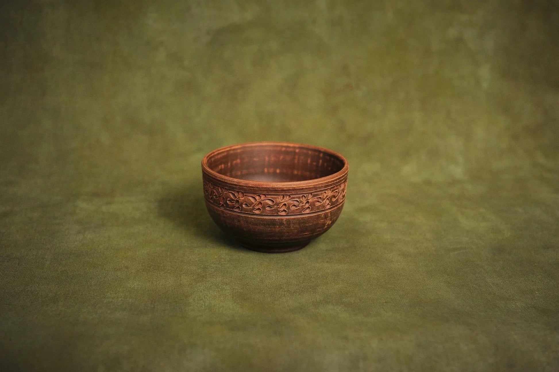 Handmade deep red clay ceramic bowl with ornate rim on a green background, Ukrainian pottery