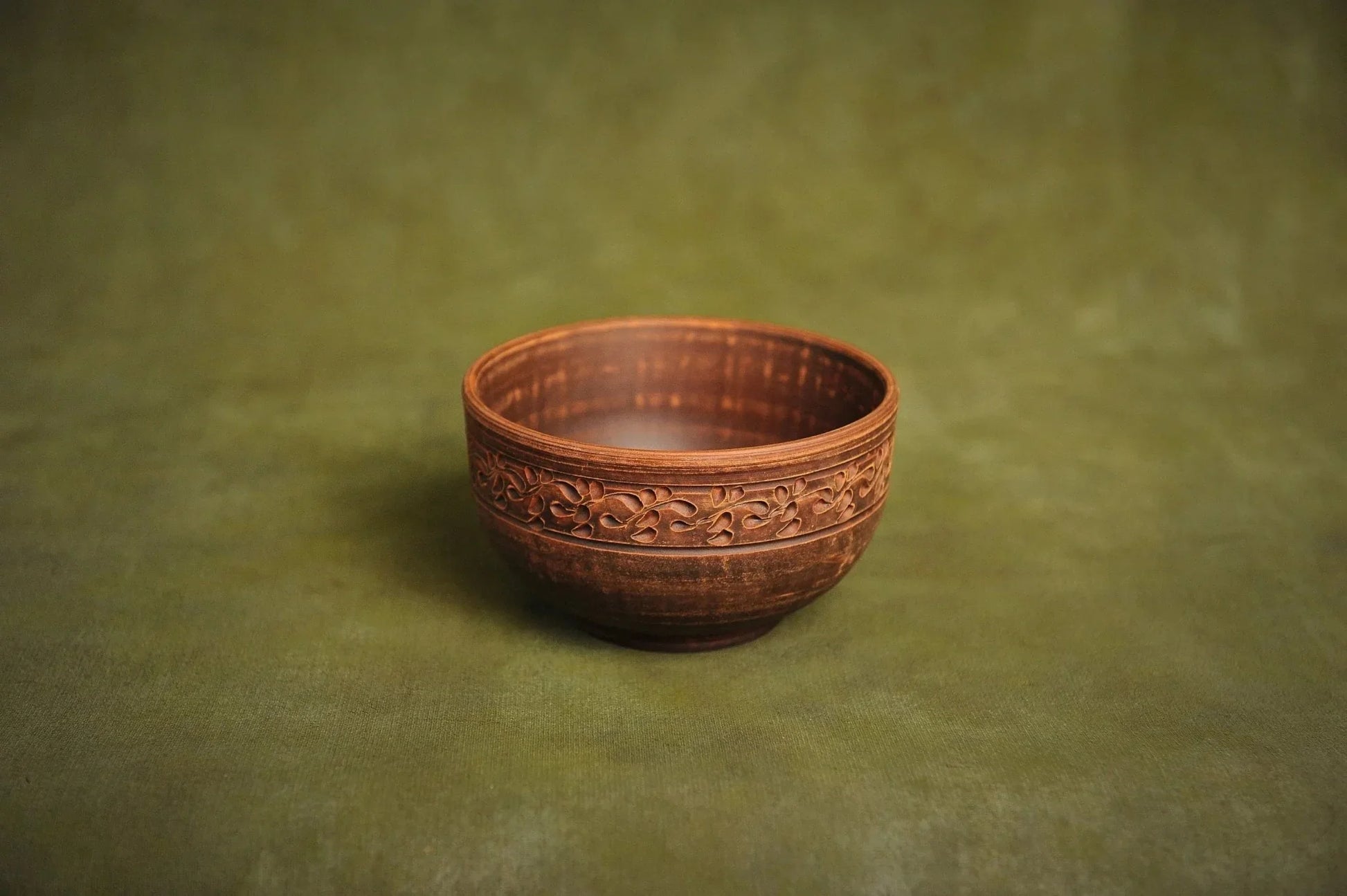 Handmade deep red clay bowl with floral carving, Ukrainian pottery on green background