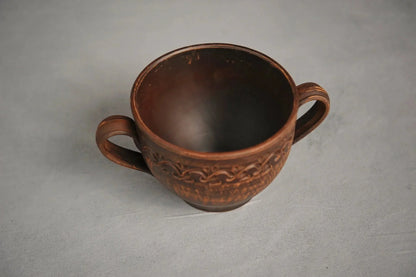 Handmade brown ceramic bowl with two handles and decorative pattern on gray background