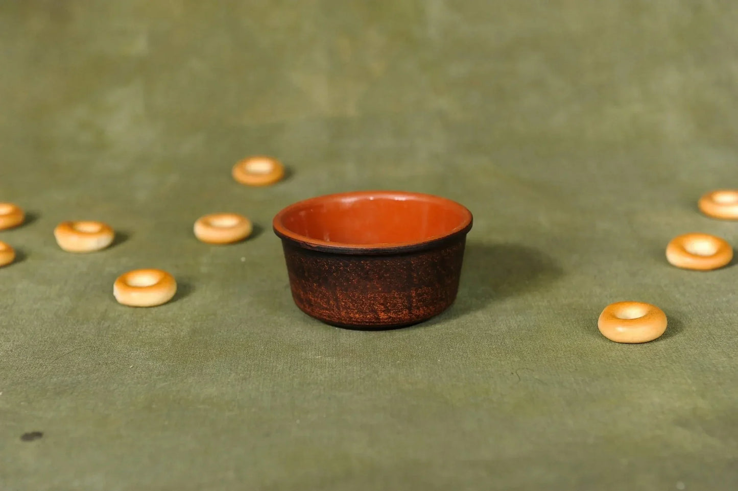 Handcrafted clay sauce bowl with brown glaze, surrounded by small round biscuits on green fabric