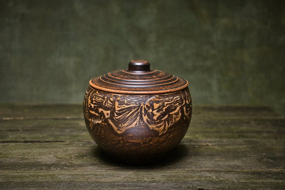 Handmade brown ceramic clay pot with lid and carved pattern on rustic wooden surface