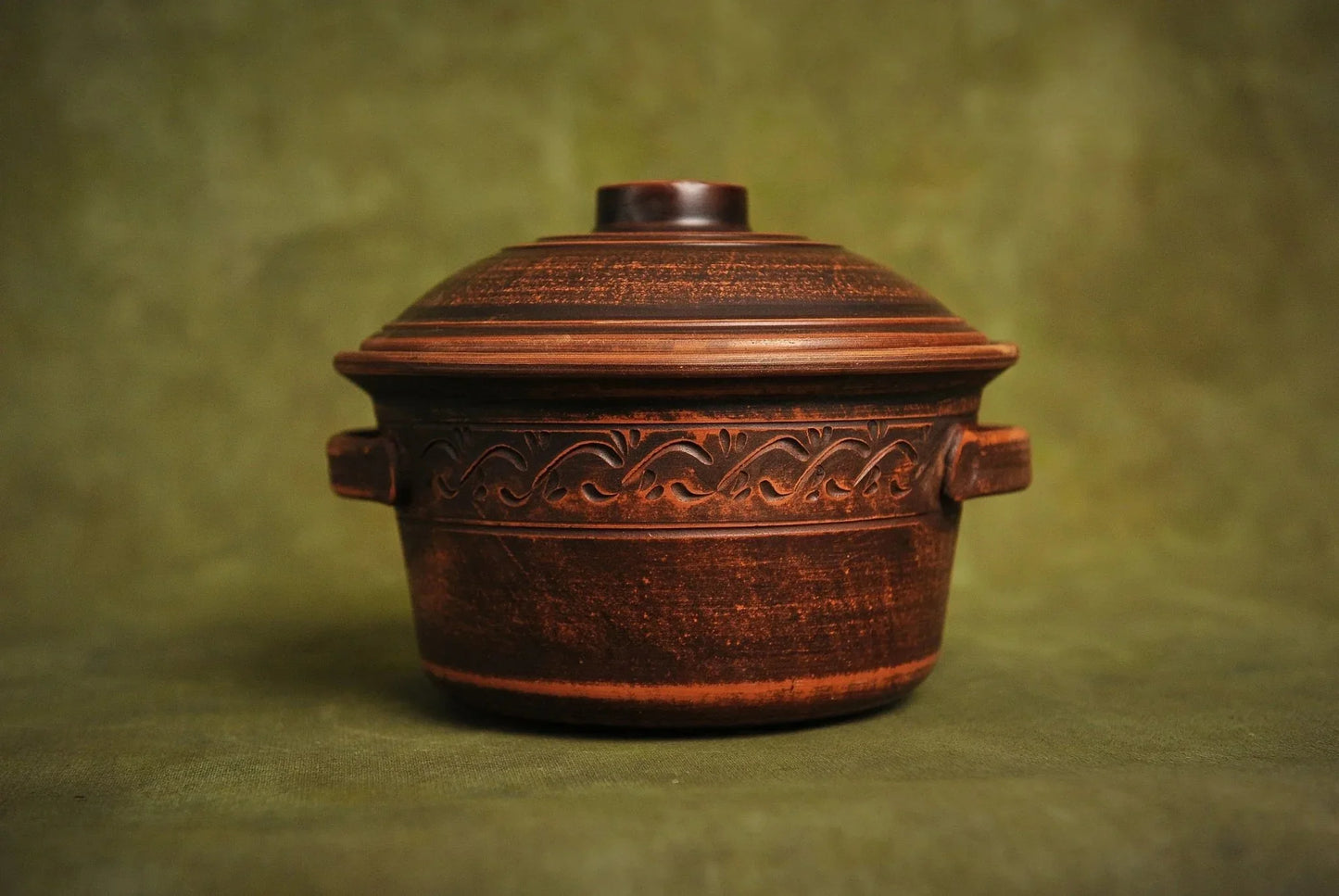 Handmade ceramic clay pot with lid, decorative handles, artisan bakeware on green background