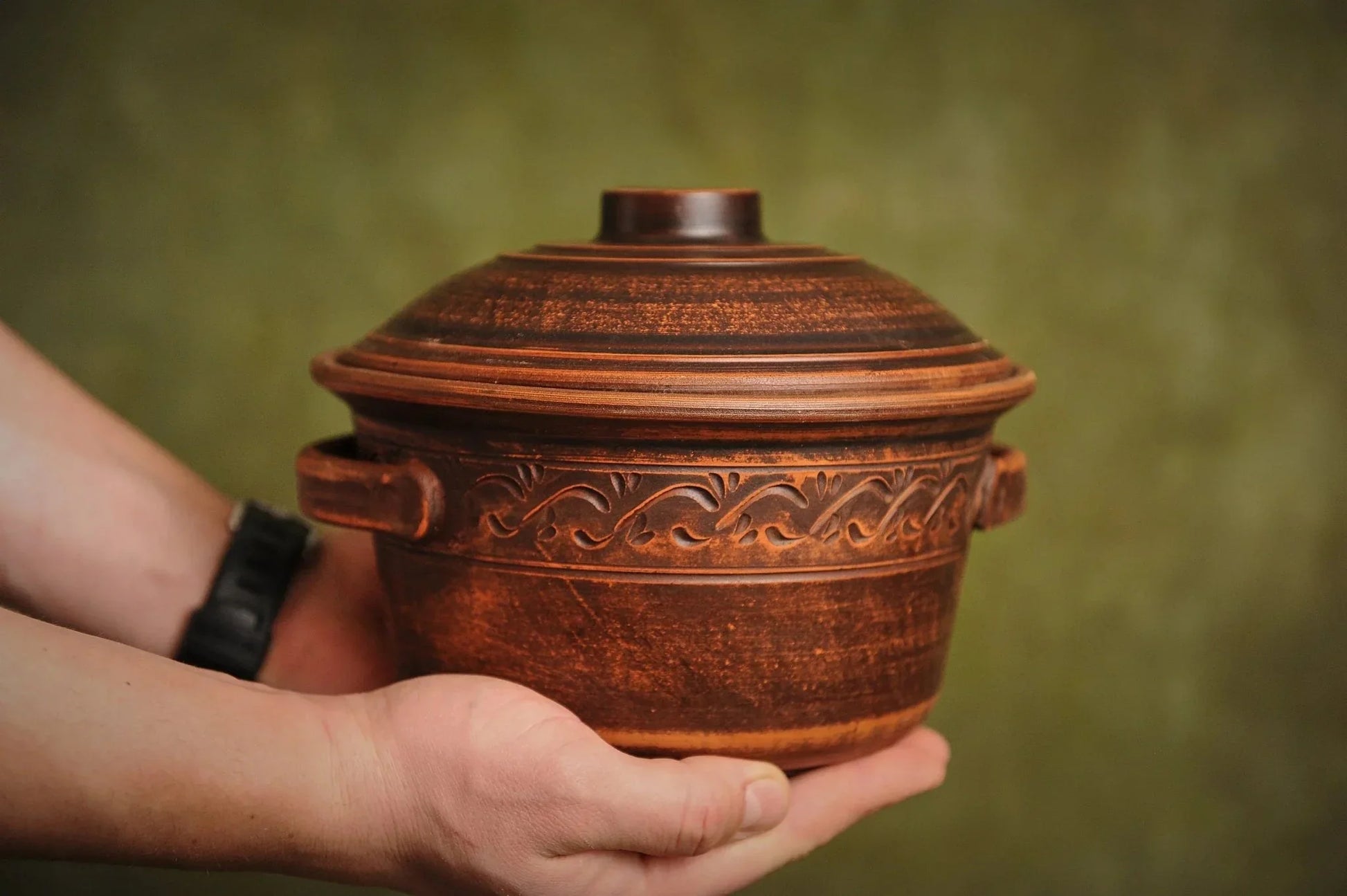 Hand holding a handmade ceramic clay pot with lid, decorative handles, and carved pattern