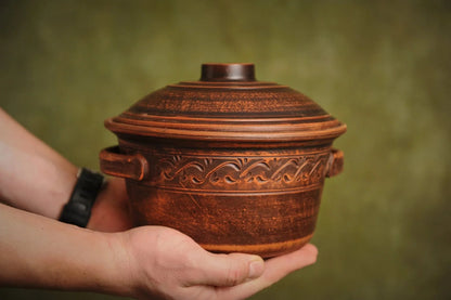 Hand holding a handmade ceramic clay pot with lid, decorative handles, and carved pattern