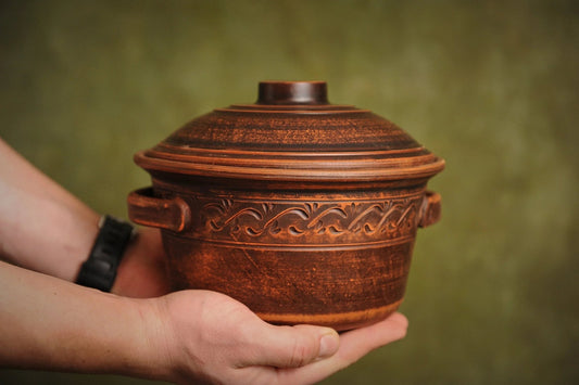 Hand holding a handmade ceramic clay pot with lid, decorative handles, and carved pattern