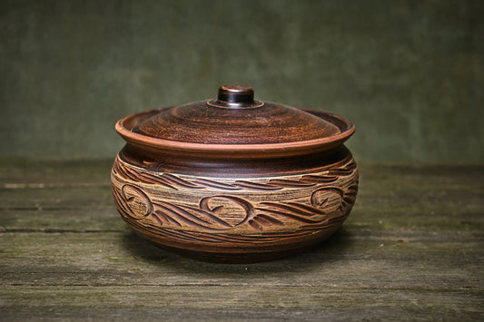 Handmade unglazed clay pot with lid and rustic carved design on wooden table