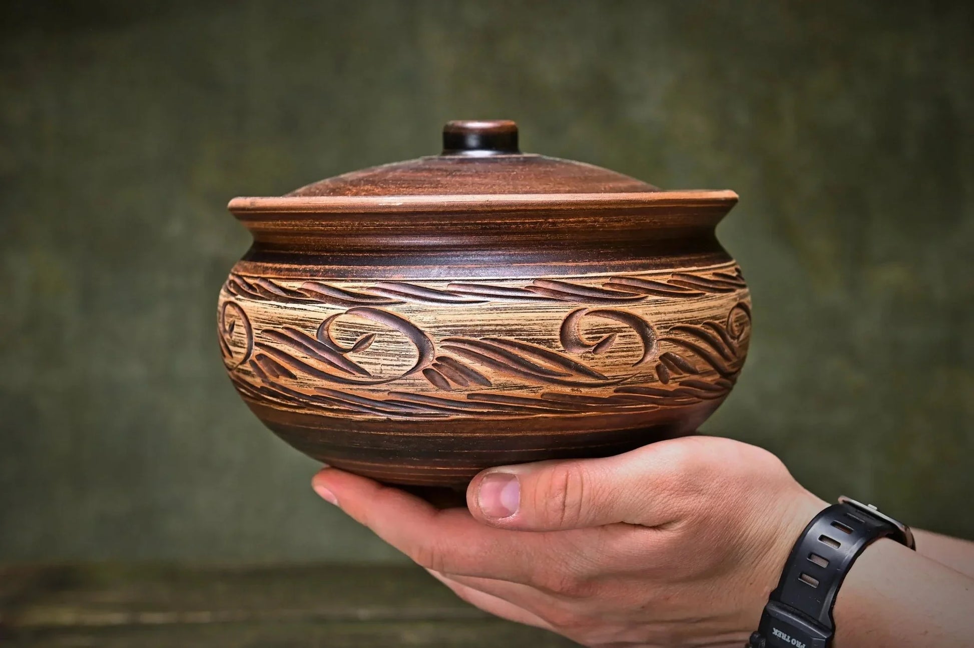 Hand holding a handmade unglazed ceramic clay pot with lid and rustic carved patterns