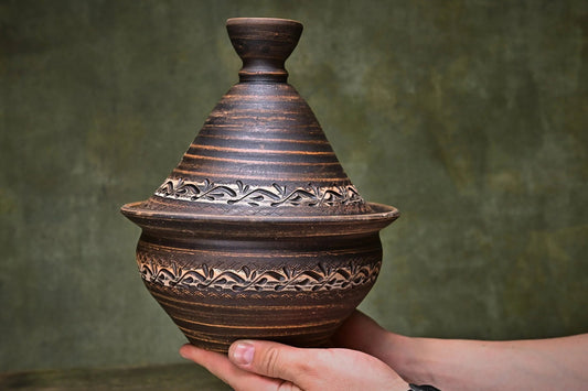 Handmade unglazed clay tagine pot with lid, rustic carved design, held in hands