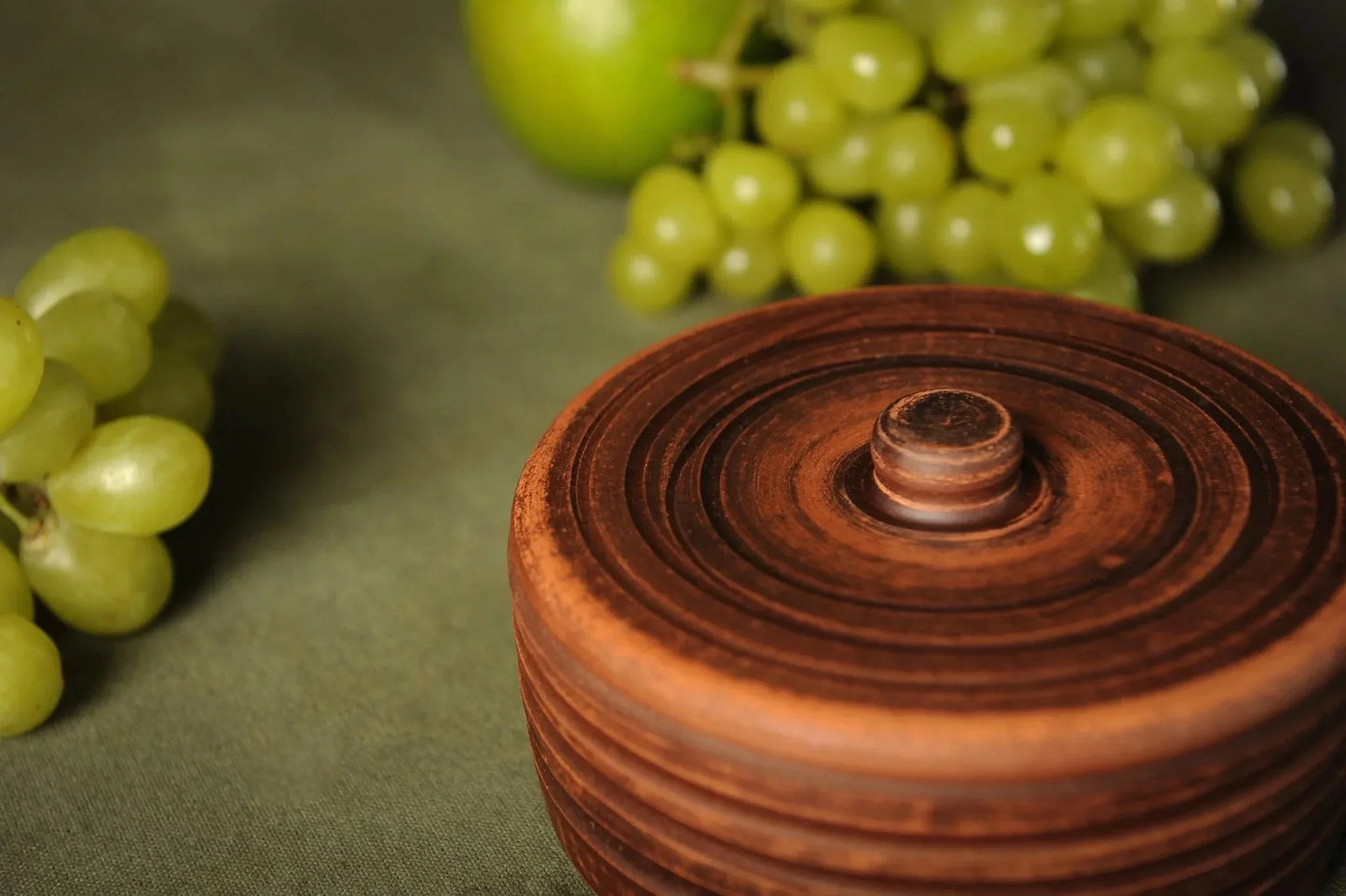 Handmade clay spice pot with lid on green fabric, grapes and apple in background