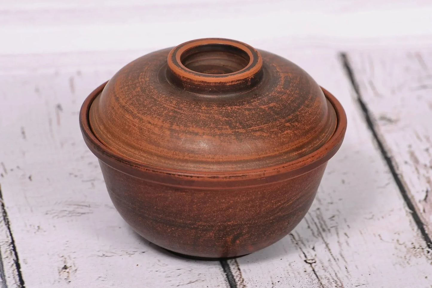 Handmade brown clay bowl with lid, rustic pottery bakeware on white wood background