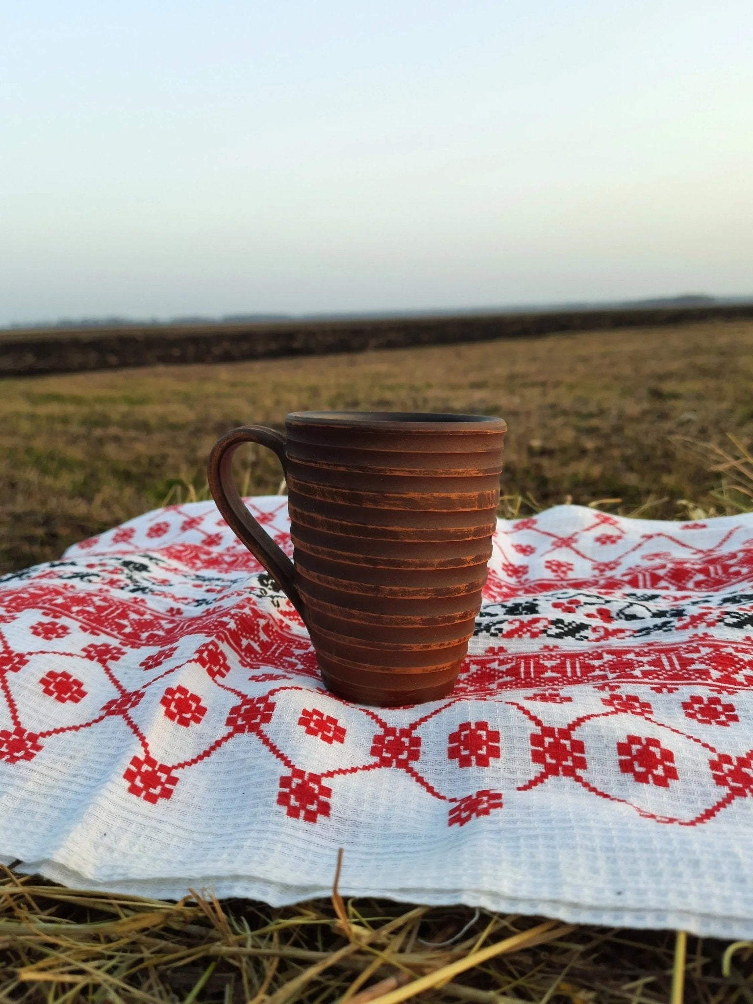 Handmade clay carved ceramic mug on embroidered cloth, outdoor setting, artisan pottery