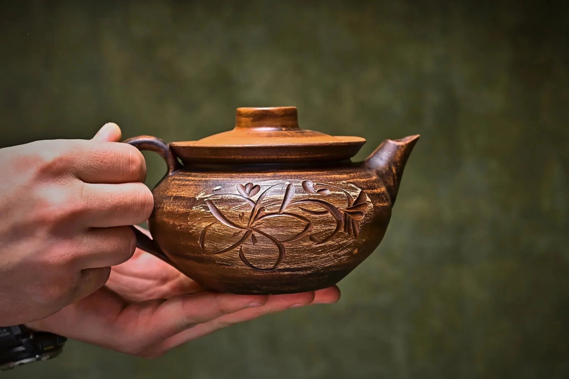 Hand holding handmade clay teapot with floral carvings, artisan home decor product