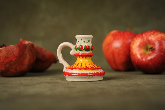 Hand-painted ceramic candlestick holder with yellow, red, and green patterns, artisan handmade, displayed with apples and pomegranate on a green background.