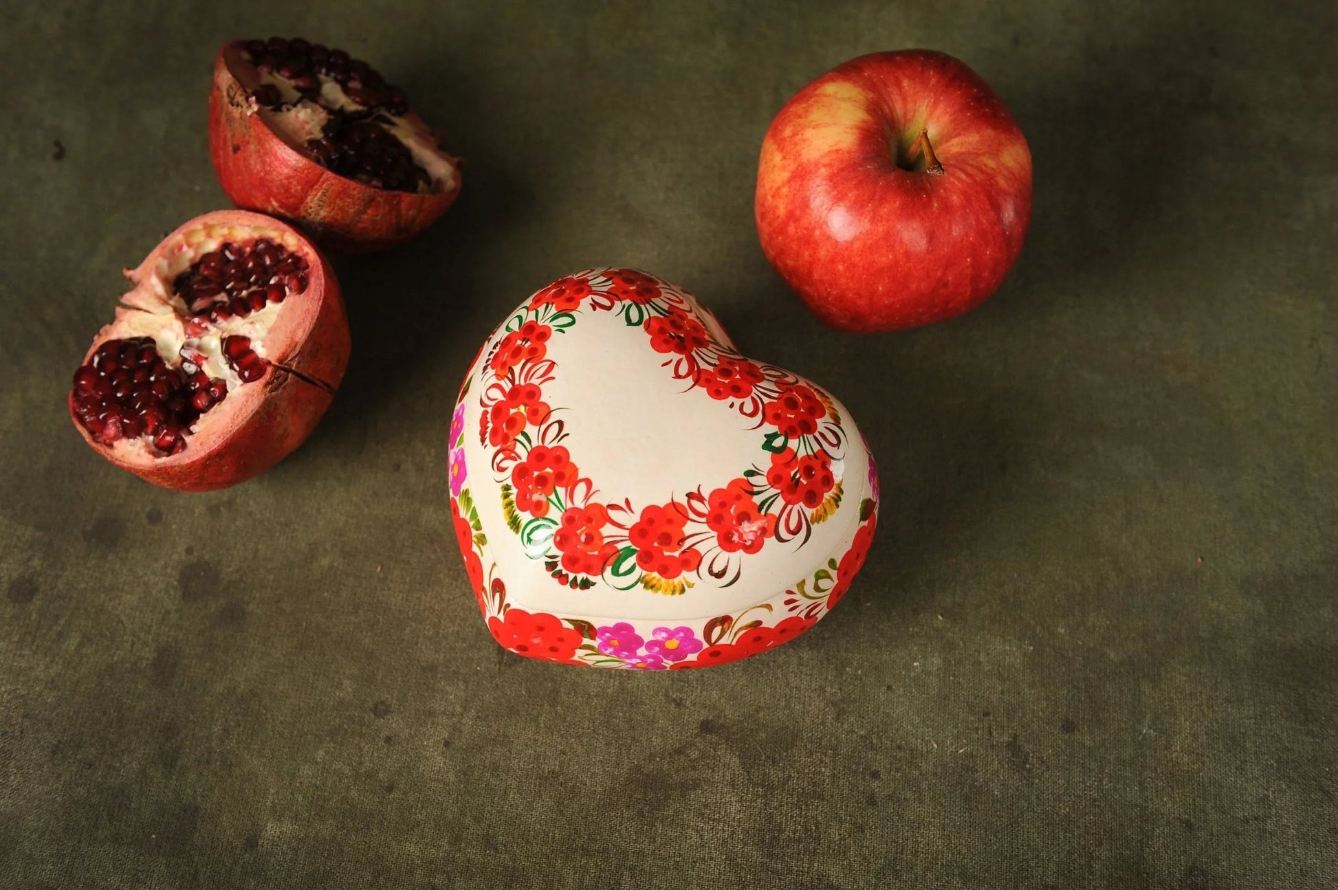 Hand-painted heart-shaped ceramic trinket box with red floral design, beside apple and pomegranate