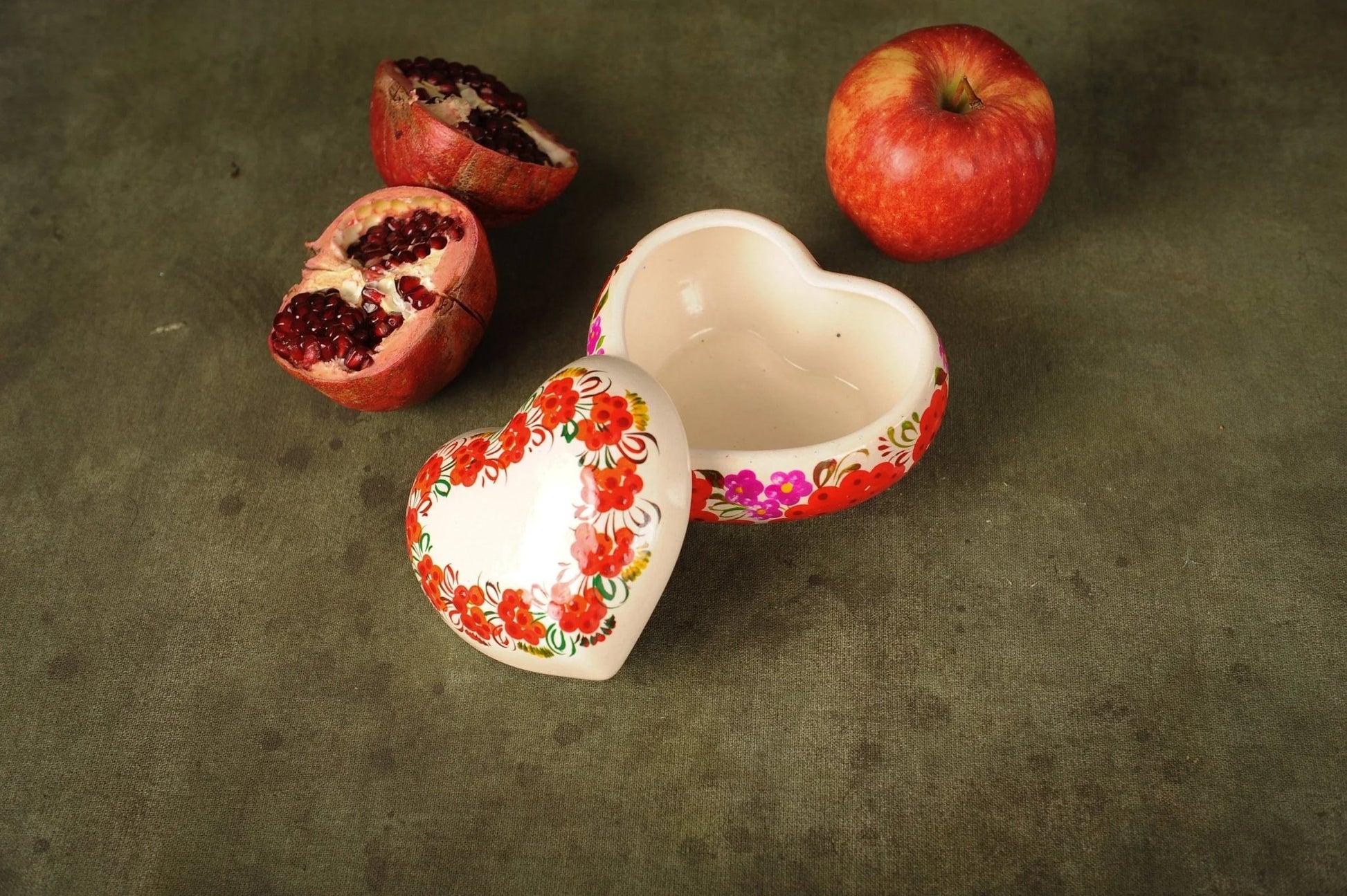 Hand-painted heart-shaped ceramic trinket box with floral design, beside pomegranate and apple