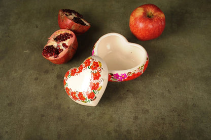 Hand-painted heart-shaped ceramic trinket box with floral design, beside pomegranate and apple