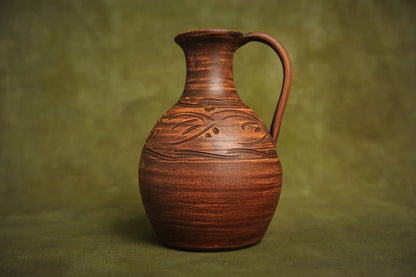 Handcrafted brown ceramic clay jug with carved details on a plain green background