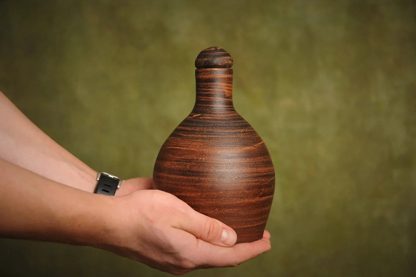 Hand holding a handmade red clay vessel jug with a rounded body and lid, rustic homeware