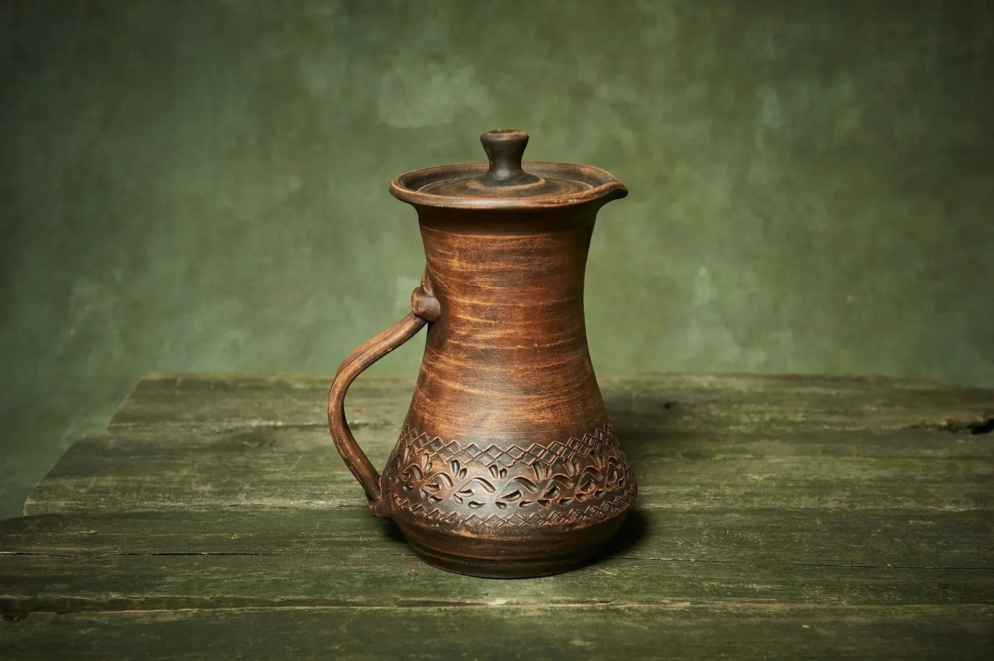Handmade ceramic pitcher with lid and ornate carvings on rustic wood table