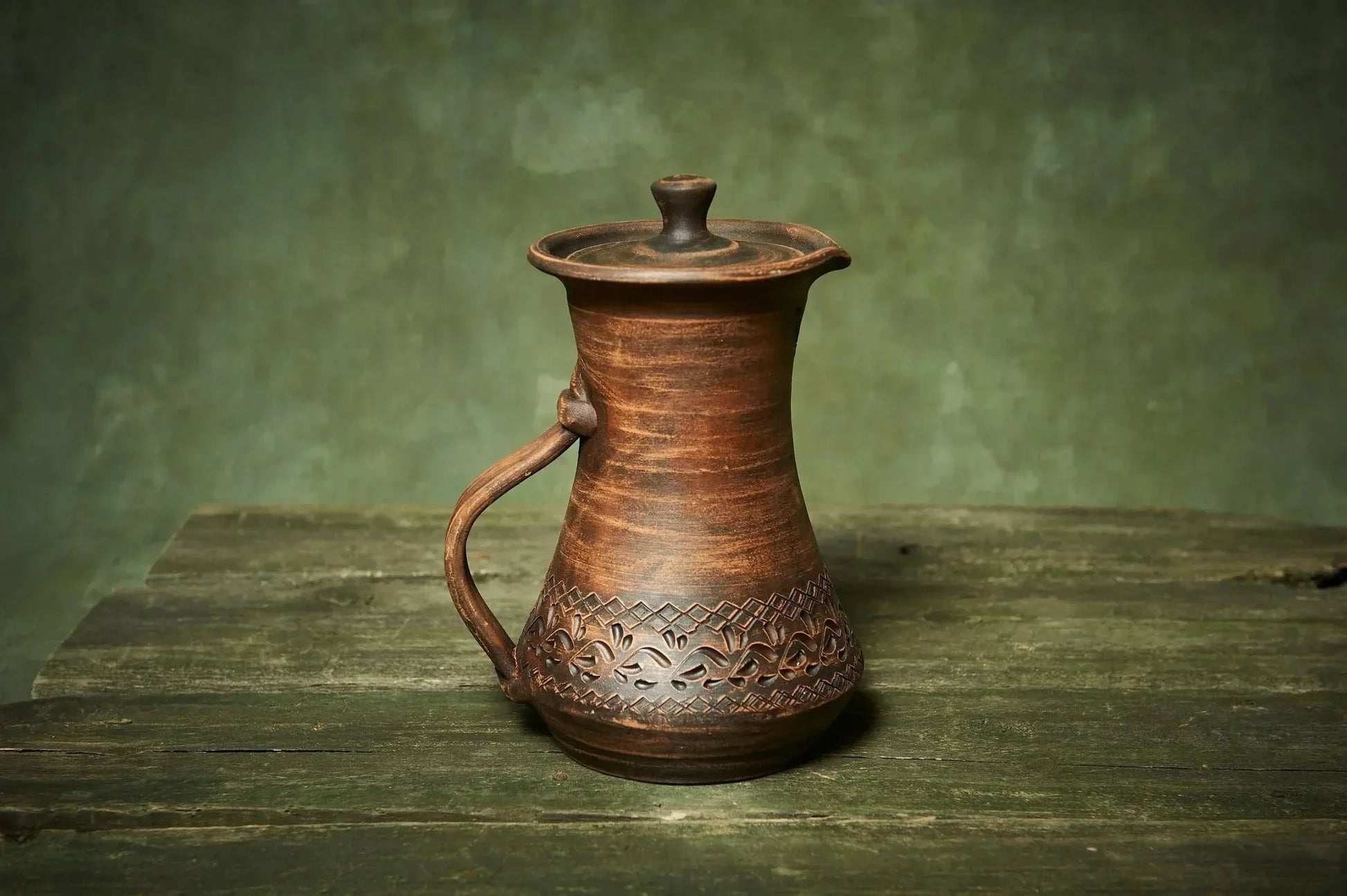 Handmade ceramic pitcher with lid and ornate carvings on rustic wood table