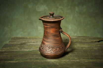 Handmade clay pitcher with lid and handle, rustic ceramic jug on wooden surface, farmhouse decor