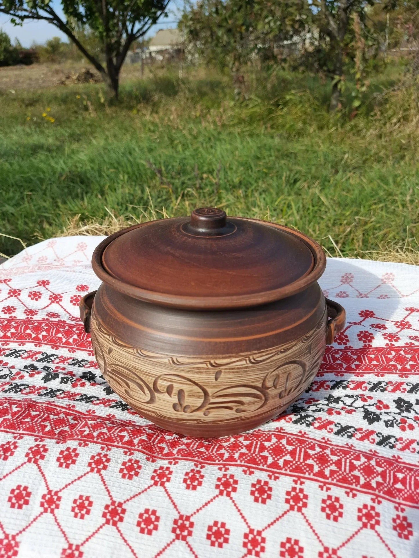 Handmade ceramic clay pot with handles and lid on patterned cloth outdoors, artisan homeware