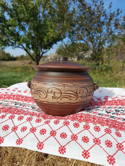 Handmade ceramic pot with lid and handles, red clay Dutch oven on embroidered cloth outdoors