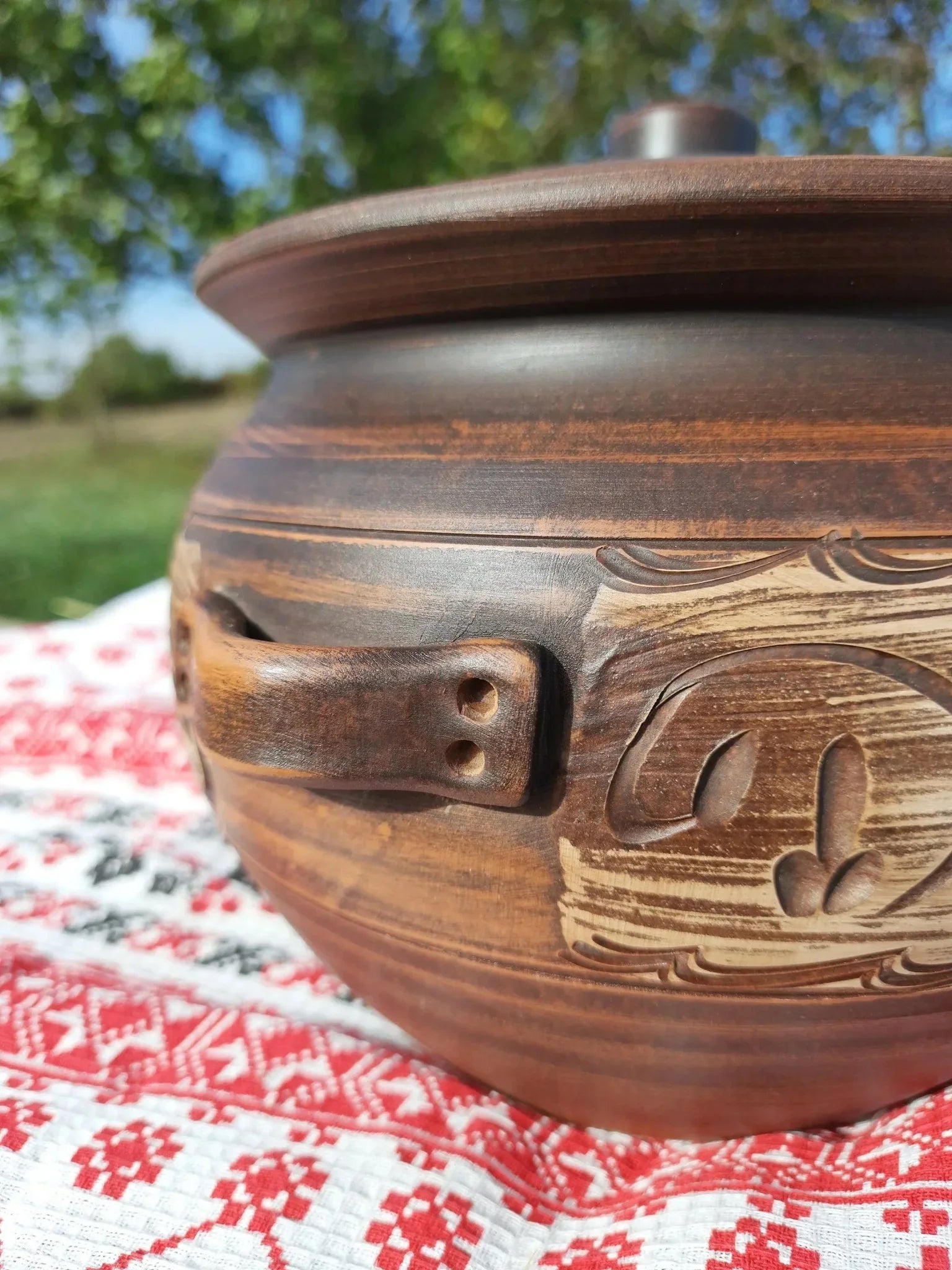 Handmade ceramic red clay pot with lid and handles, artisan cookware on embroidered cloth, outdoor setting