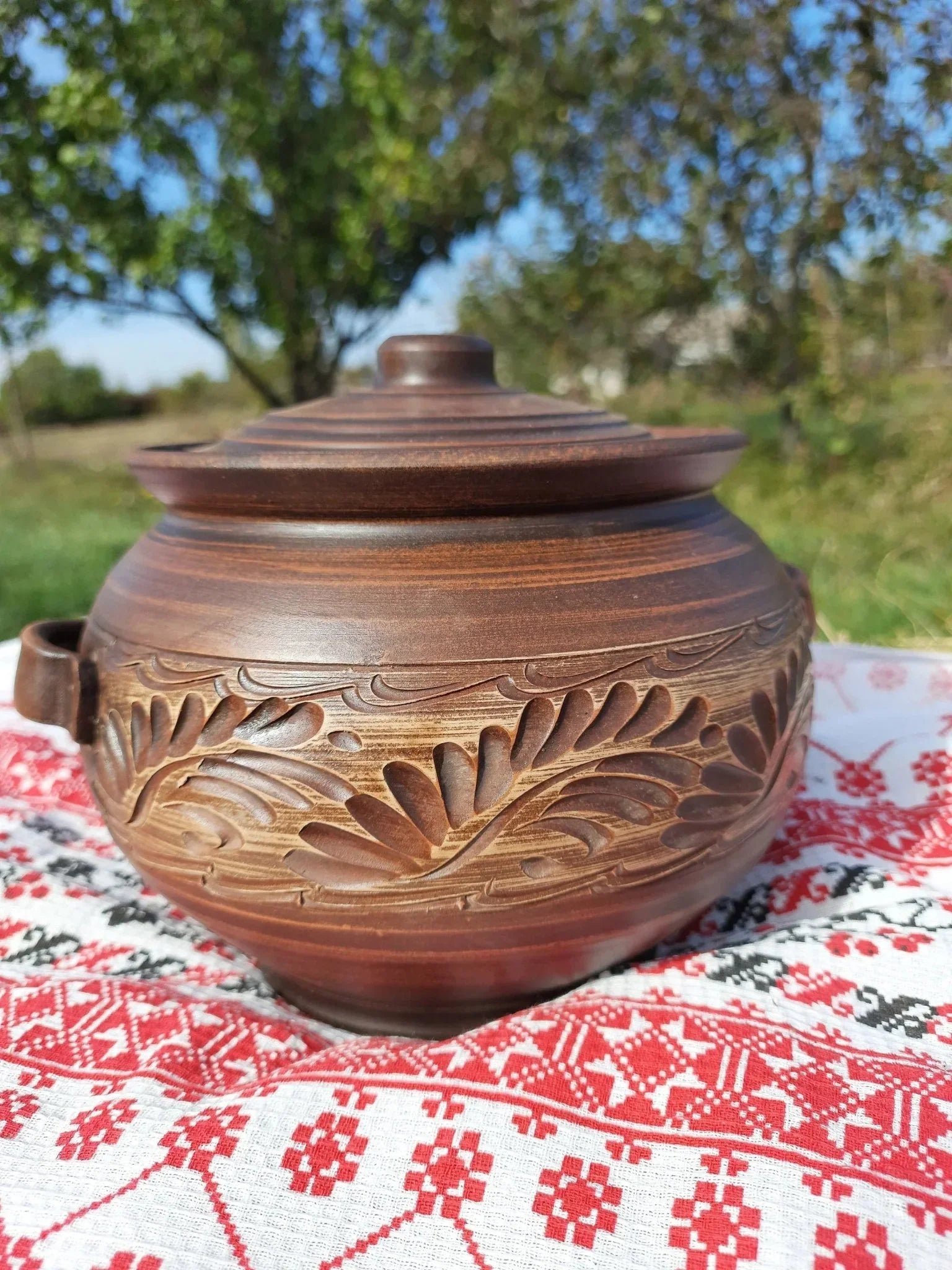 Handmade ceramic pot with handles and lid, featuring carved red clay, displayed outdoors on an embroidered cloth.