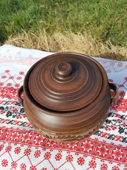 Handmade ceramic pot with lid and handles, red clay, on embroidered cloth outdoors
