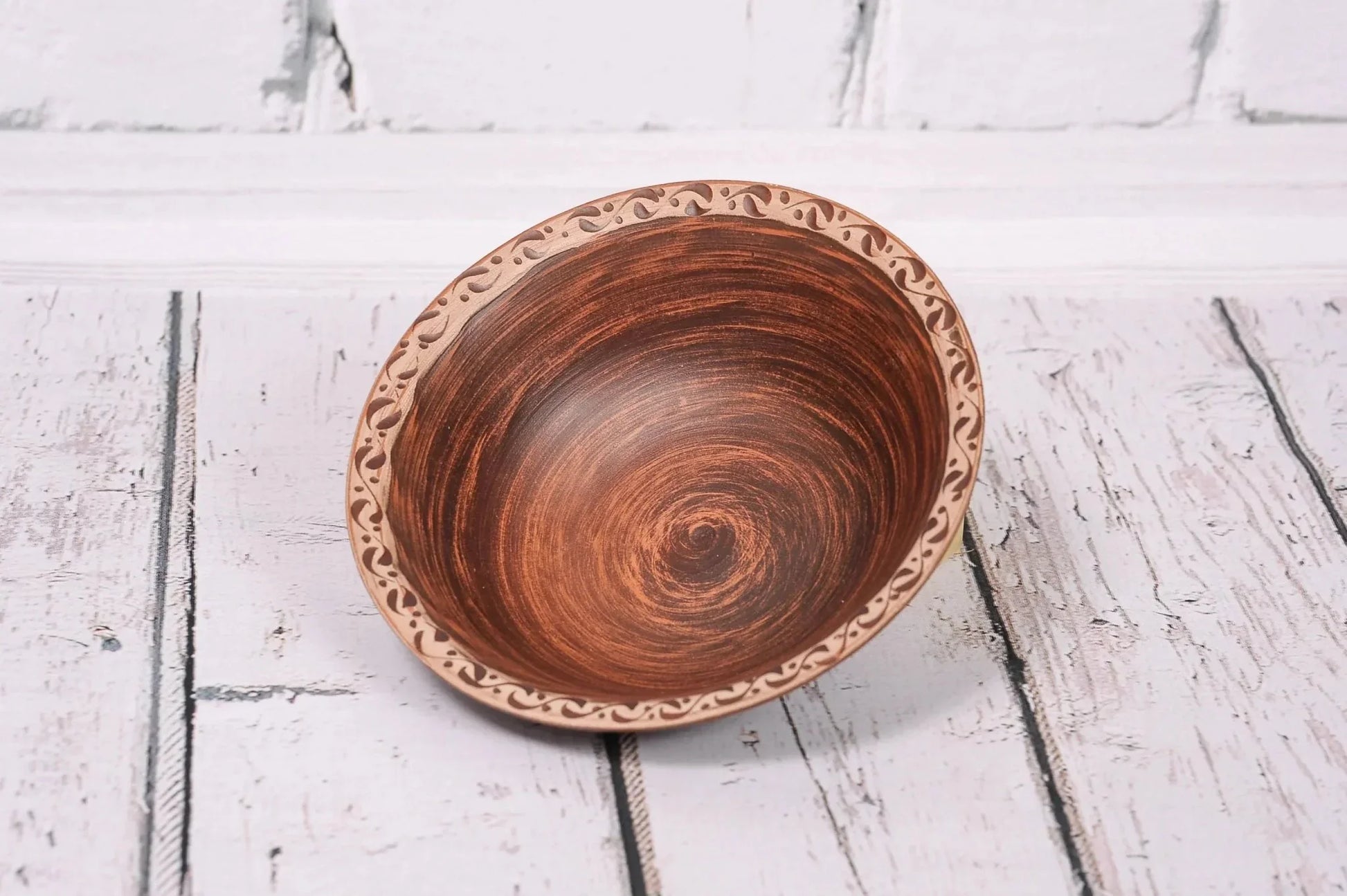 Handmade clay bowl with decorative rim, rustic ceramic pottery on white wooden surface