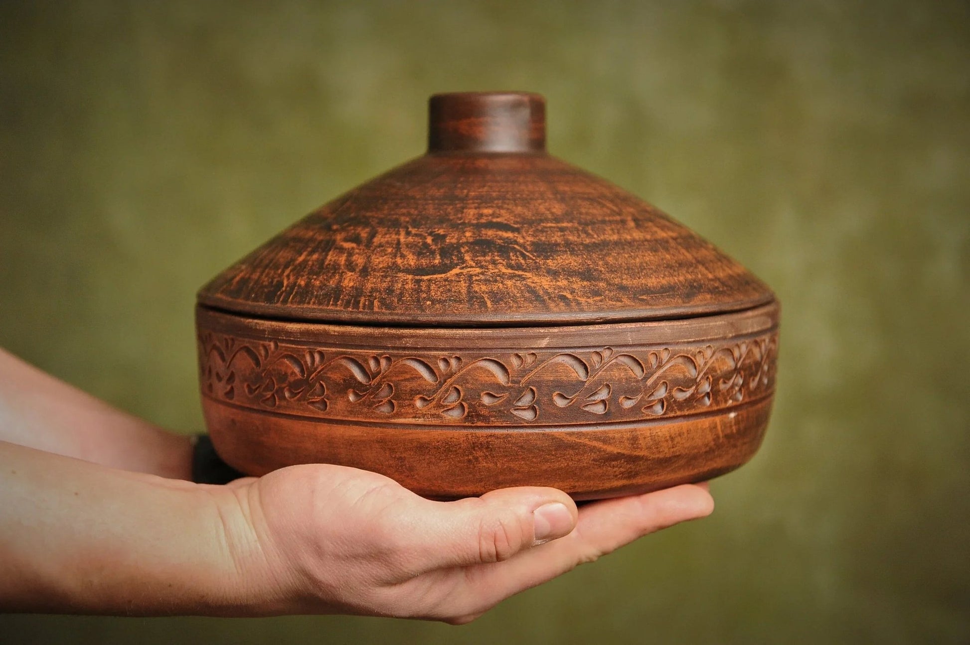 Hand holding a handmade clay cooking pot with lid, featuring ornate carved patterns, organic ceramics cookware