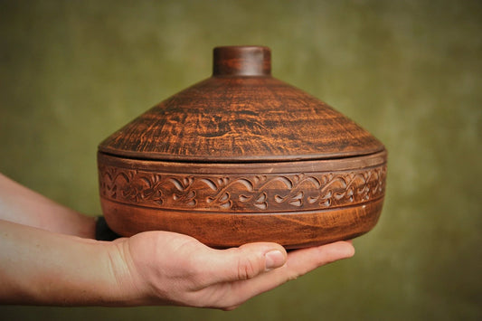 Hand holding a handmade clay cooking pot with lid, featuring ornate carved patterns, organic ceramics cookware