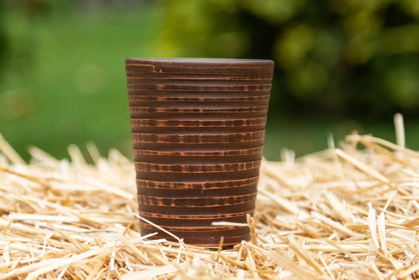 Handmade clay pottery tumbler with horizontal stripes on straw outdoors, artisan drinkware