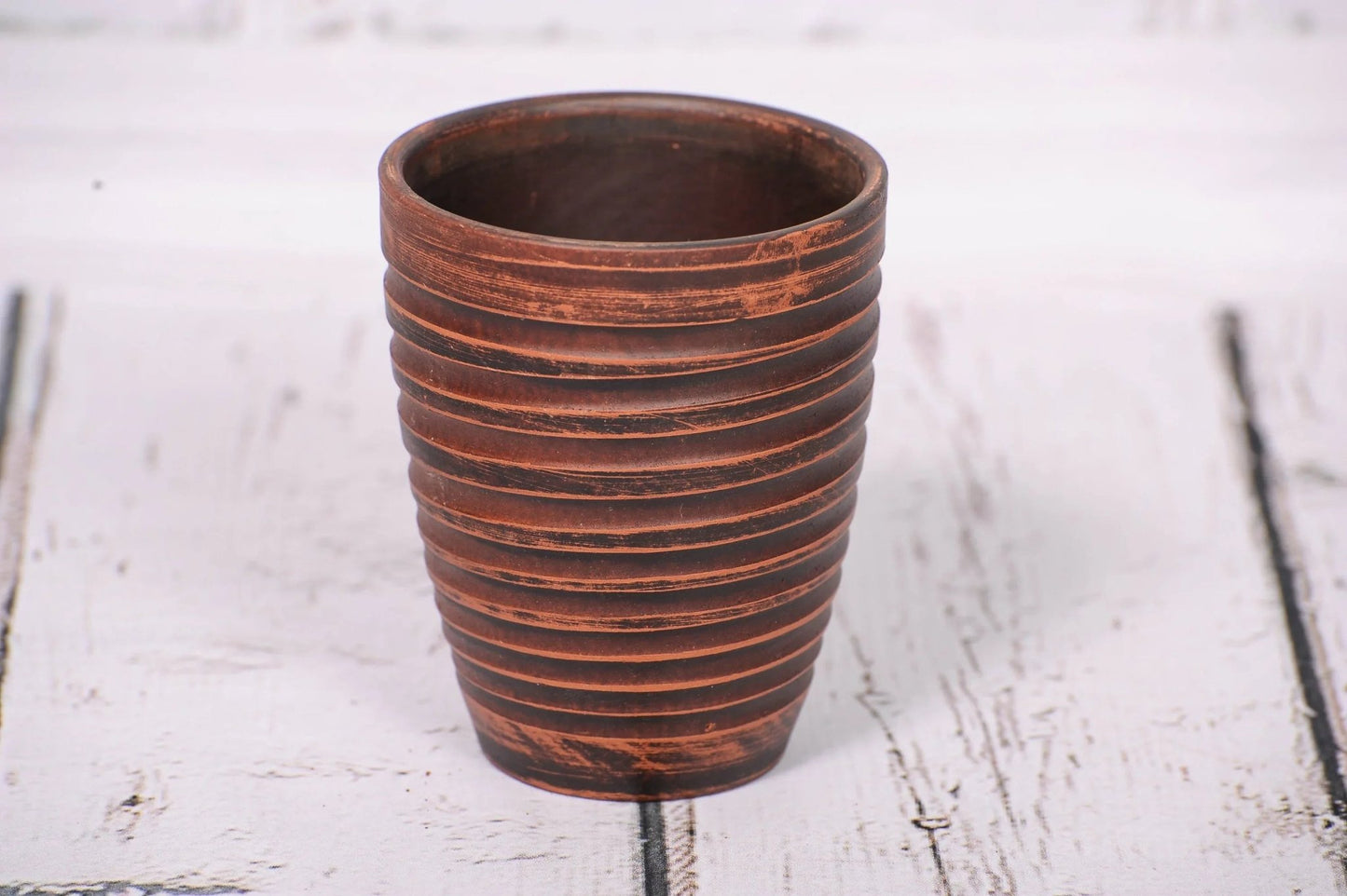 Handmade clay tumbler with textured stripes on rustic white wood, artisan pottery cup