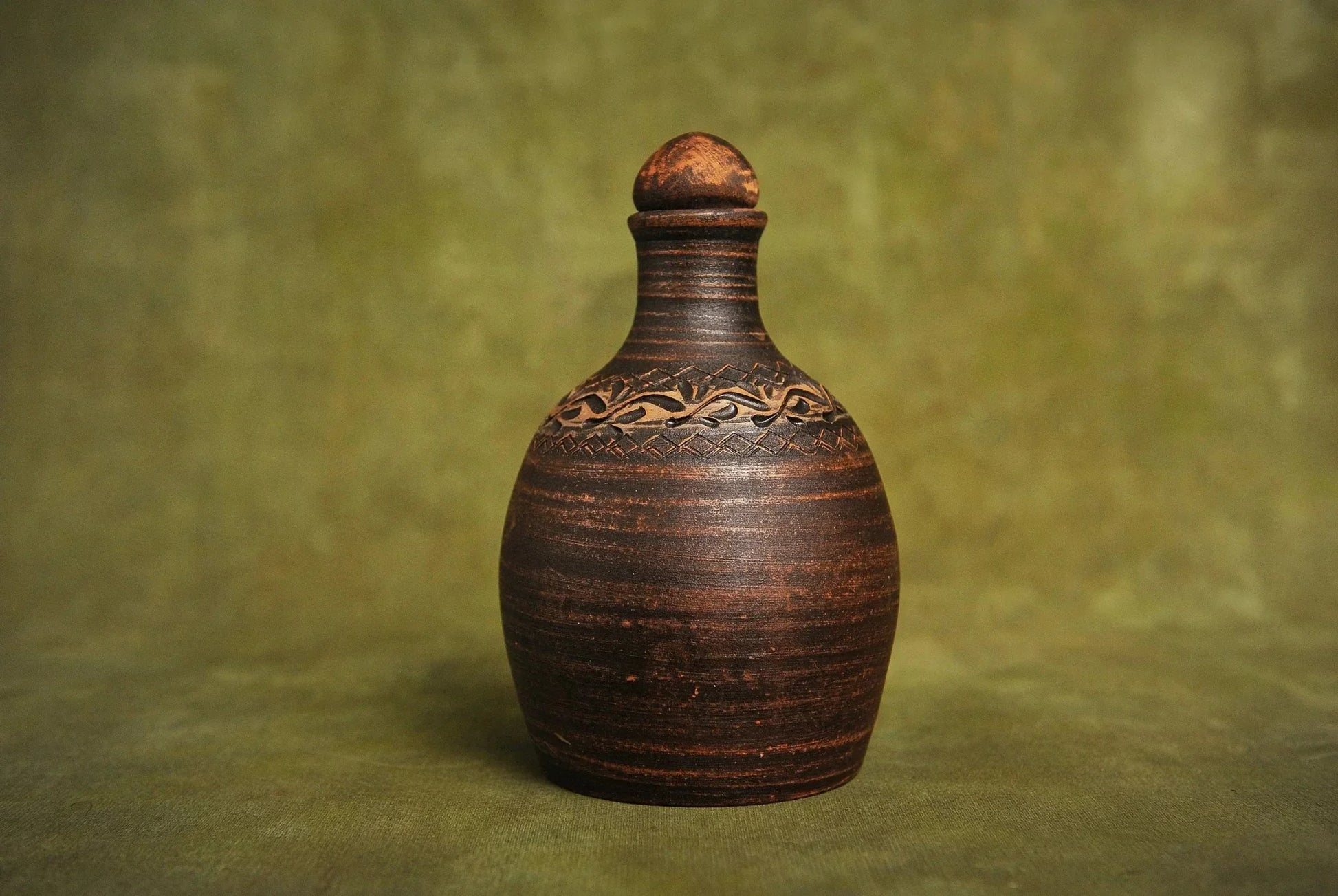 Handmade red clay bottle with carved decorative pattern, rustic homeware on green background