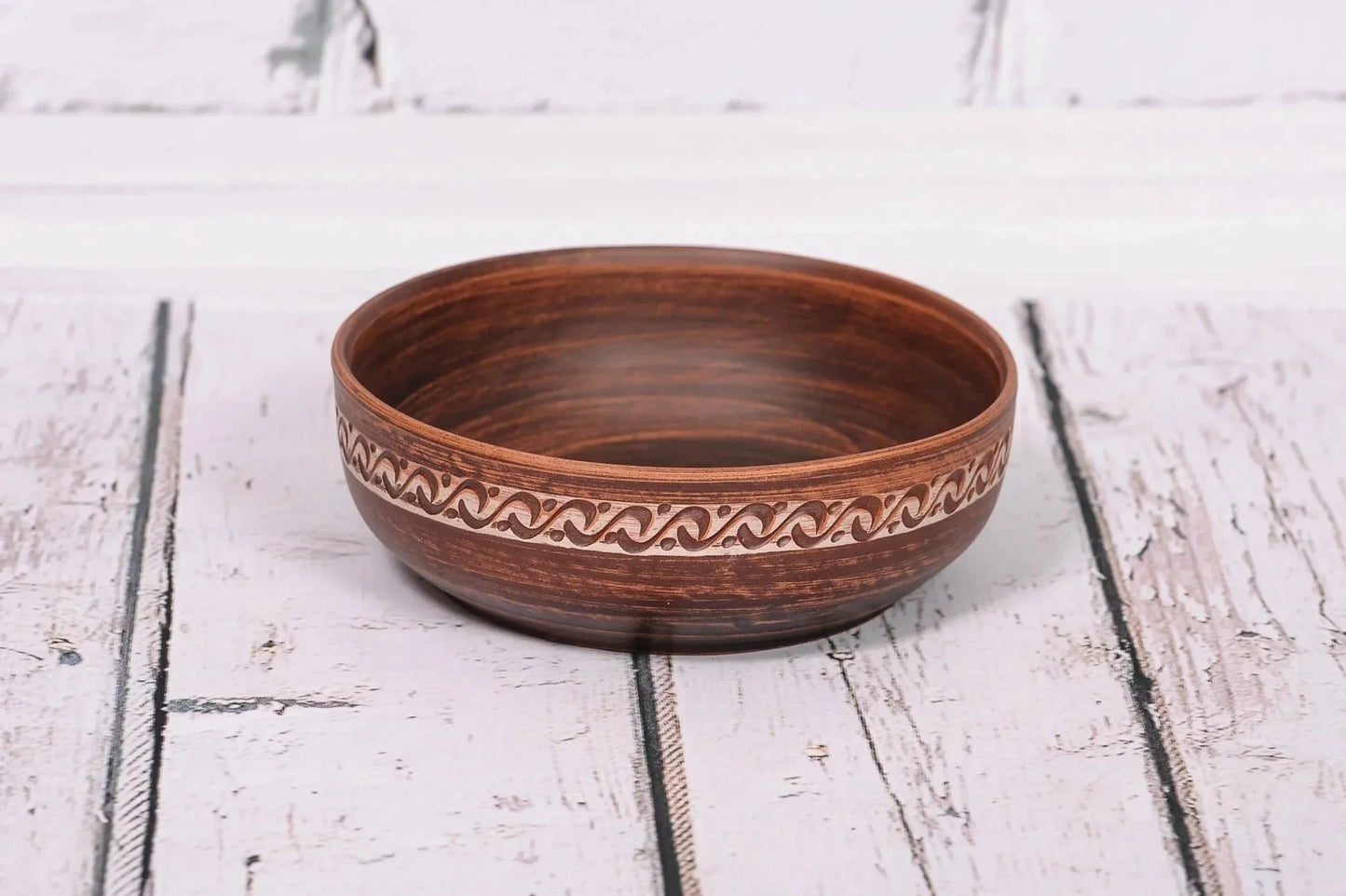 Handmade red clay bowl with decorative rim, rustic ceramic pottery on white wooden surface
