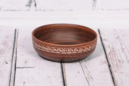 Handmade red clay bowl with decorative rim, rustic ceramic pottery on white wooden surface