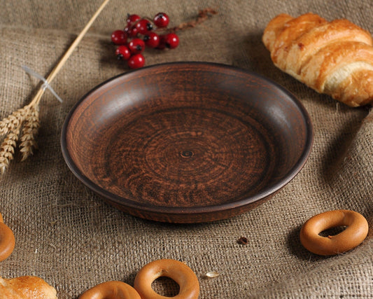 Handmade brown clay plate on burlap with bread, wheat, berries—artisan ceramic homeware