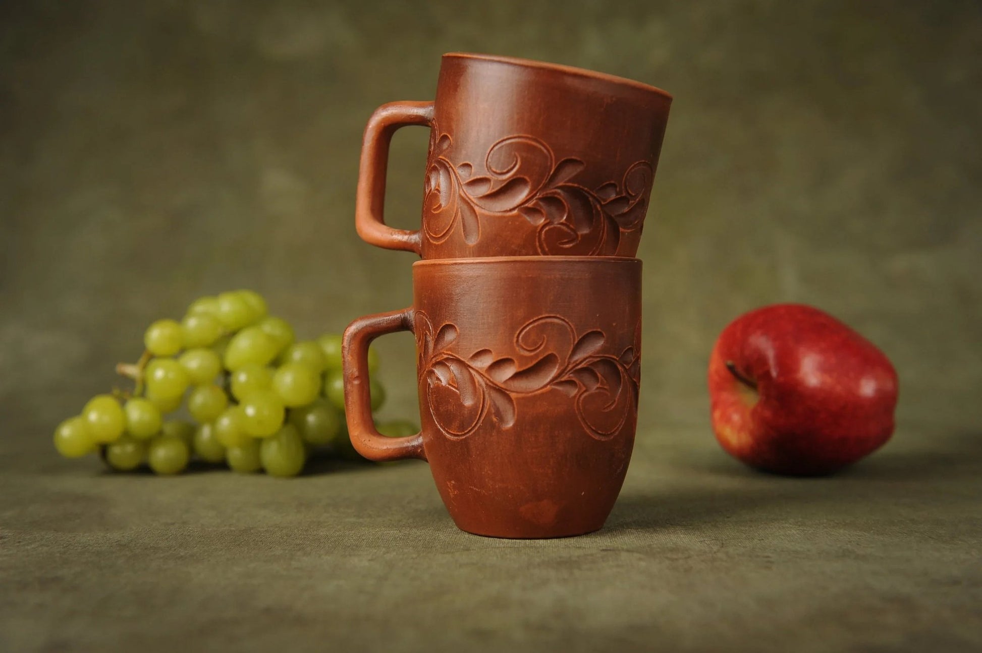 Set of two handmade red clay ceramic cups with floral carvings, displayed with grapes and an apple.