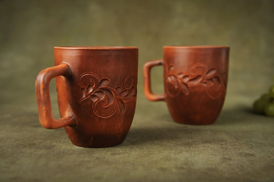 Set of two handmade red clay ceramic cups with floral carvings on green background