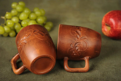 Set of two handmade clay ceramic cups with carved floral design, on fabric with grapes and apple.