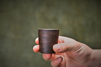 Hand holding a handmade ceramic shot glass with a rustic brown clay finish