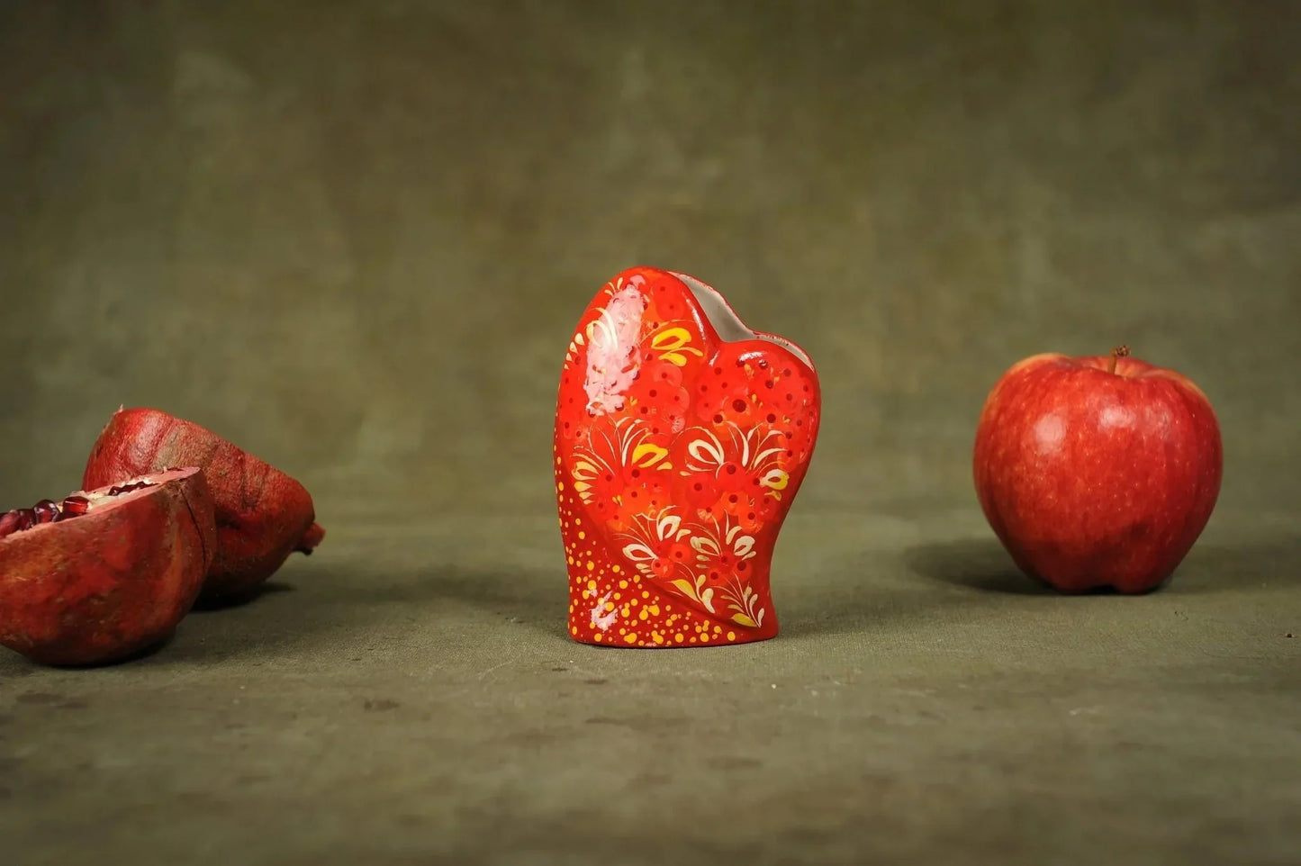 Hand-painted red ceramic heart-shaped vase with floral patterns, displayed with apple and pomegranate.