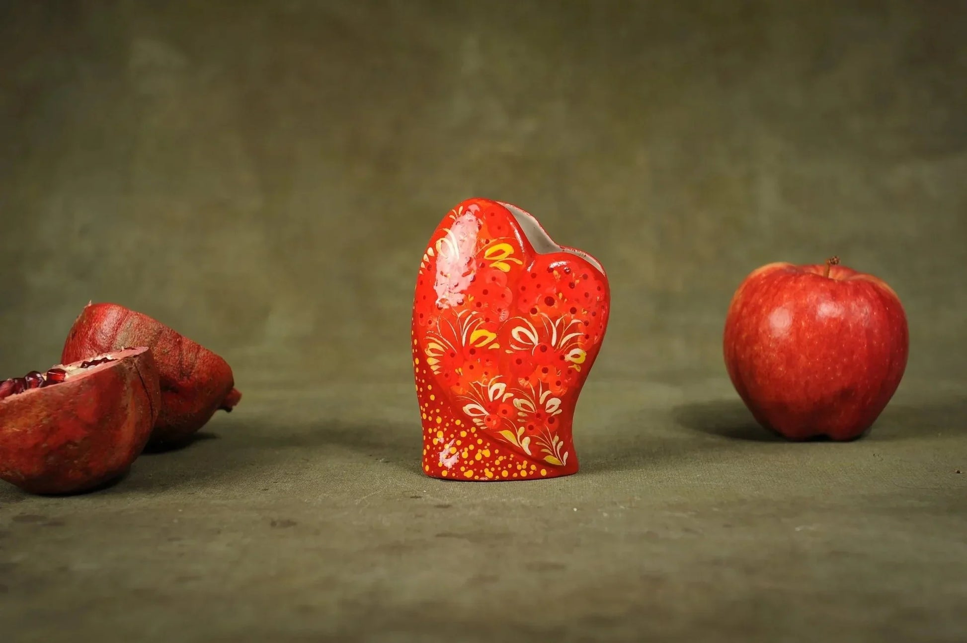 Hand-painted red ceramic heart-shaped vase with floral patterns, displayed with apple and pomegranate.