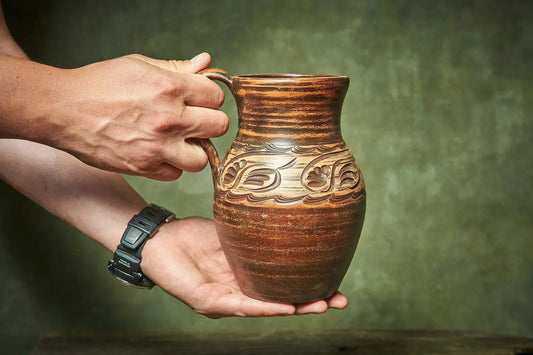 Hand holding a rustic handmade clay pitcher with carved floral design, farmhouse pottery
