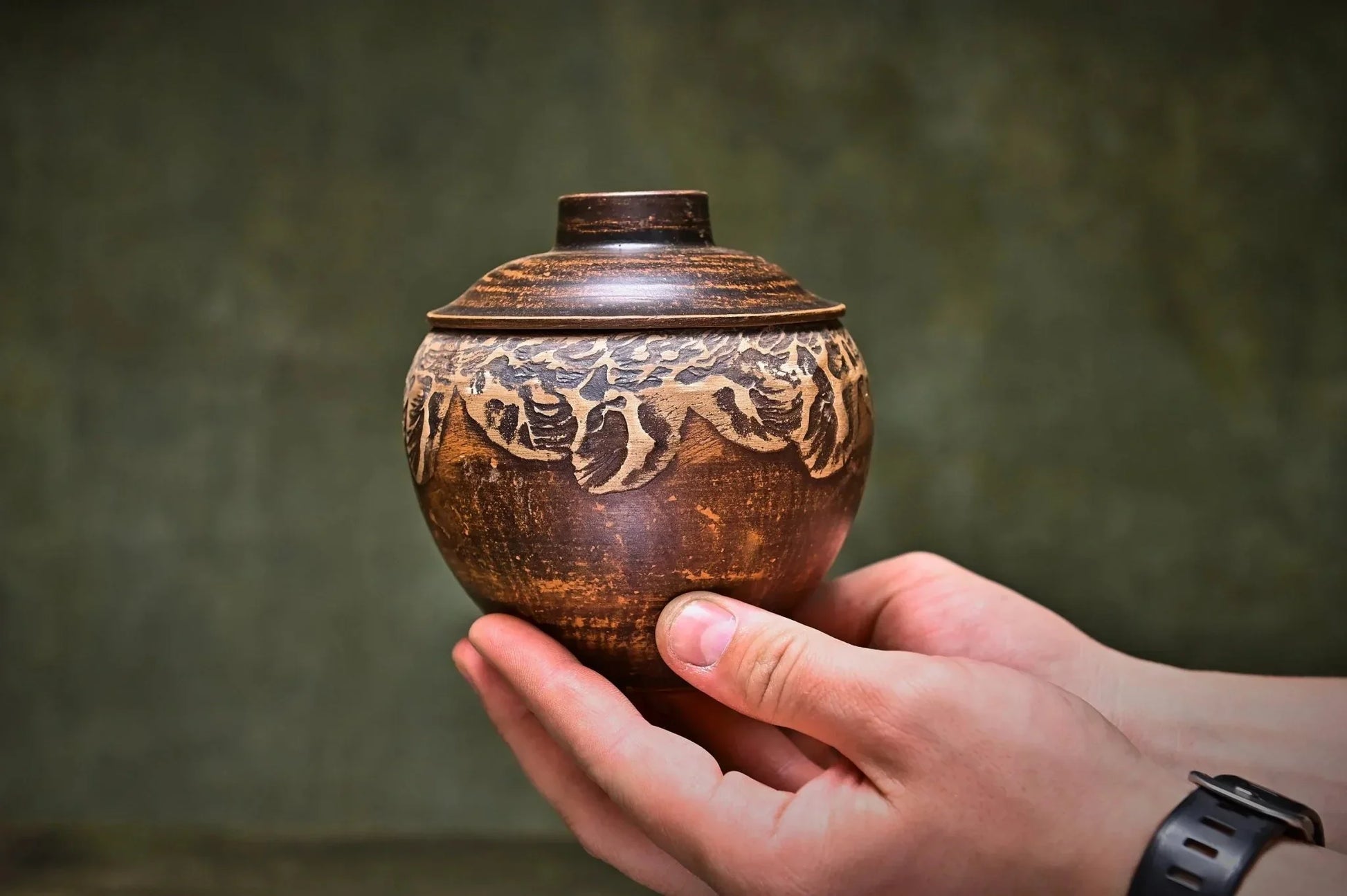 Hand holding a small handmade Ukrainian ceramic pot with lid, rustic carved design