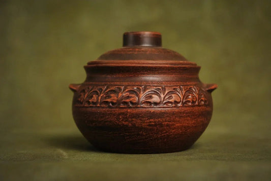 Handmade small brown ceramic pot with lid and ornate leaf pattern, on green background